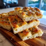 Close-up of stacked slices of airy rosemary focaccia bread with coarse salt on a wooden board.