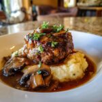 A close-up of a savory Salisbury steak served over creamy mashed potatoes, topped with mushroom gravy and fresh herbs.