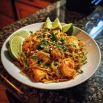 A close-up of a vibrant plate of shrimp Pad Thai, garnished with fresh lime wedges, chopped peanuts, and cilantro.