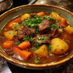 A close-up of a bowl filled with tender slow cooker beef stew, featuring chunks of beef, potatoes, carrots, and fresh parsley garnish.