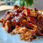 Close-up of tender slow cooker pulled pork coated in a rich BBQ sauce, served on a light blue plate.