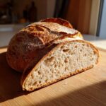 A golden-brown, crusty loaf of sourdough bread with one slice cut, revealing a soft, airy interior.