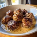 A close-up of golden-brown Swedish meatballs swimming in a rich, creamy sauce with visible herbs.