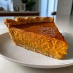 A close-up view of a perfect slice of sweet potato pie on a white plate, showcasing its golden crust and smooth filling.