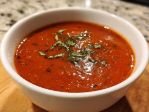 A close-up of a white bowl filled with rich, red Tomato Basil Soup, garnished with fresh basil leaves.