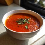 A close-up of a white bowl filled with vibrant red tomato basil soup, garnished with fresh basil leaves and black pepper.