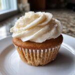 A single, perfectly baked vanilla cupcake topped with swirls of creamy white frosting on a white plate.