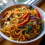 A close-up of a bowl filled with vibrant Vegetable Lo Mein, featuring noodles, red bell peppers, bok choy, and onions.