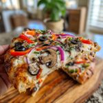 A hand holding a slice of loaded veggie pizza, showcasing mushrooms, red peppers, olives, and red onion.