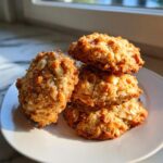A stack of four golden brown almond joy cookies, studded with visible nuts, sitting on a white plate near a window.