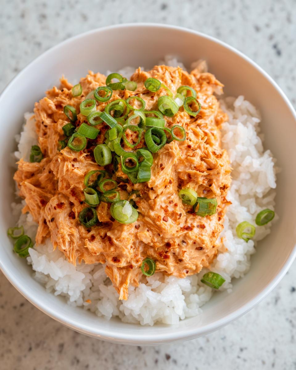A close-up of a white bowl filled with white rice topped with creamy, spicy tuna rice mixture and sliced green onions.