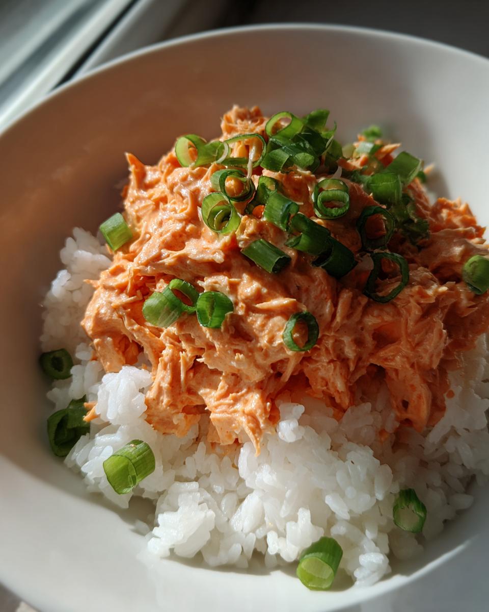 A close-up of a bowl of white rice topped with creamy, orange-colored tuna rice mixture and fresh green scallions.