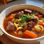 Close-up of a steaming bowl of rich beef noodle soup with chunks of beef, carrots, celery, and noodles, topped with parsley.