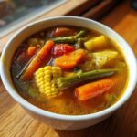 Close-up of a white bowl filled with colorful, chunky vegetable soup, featuring carrots, corn, and green beans.