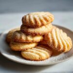 A close-up stack of golden, ridged butter cookie treats generously sprinkled with granulated sugar on a light plate.