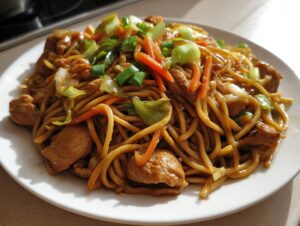 Close-up of a plate featuring a generous serving of chicken chow mein recipe with noodles, chicken pieces, carrots, and green onions.