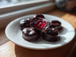 A plate of homemade chocolate covered cherries, one cut in half showing the bright red cherry filling inside the dark chocolate shell.