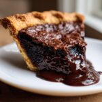 A close-up of a rich, dark slice of hot fudge pie with a gooey chocolate sauce dripping onto a white plate.