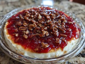 A round serving of pepper jelly cheese dip topped with bright red jelly and candied pecans in a clear glass dish.