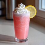 A tall glass of frosty pink lemonade topped with whipped cream and a lemon slice garnish.