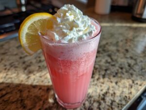 A tall glass of bright pink lemonade topped with whipped cream and garnished with a lemon slice.