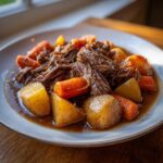 A close-up of tender, shredded pot roast served with large chunks of carrots and potatoes in rich gravy.
