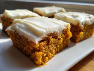 Four moist, square pumpkin bars with thick, white cream cheese frosting served on a white plate.