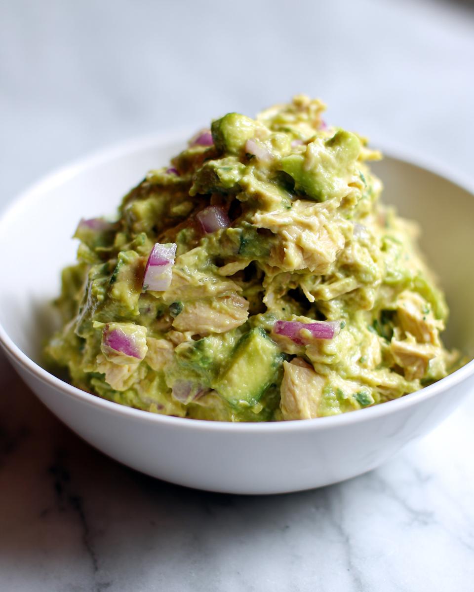 A close-up of creamy avocado chicken salad mixed with chunks of avocado and diced red onion, served in a white bowl.