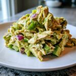 A close-up mound of creamy avocado chicken salad mixed with chunks of green pepper and red onion on a white plate.