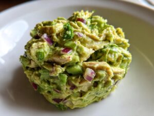 Close-up of a scoop of creamy avocado chicken salad mixed with red onion and celery on a white plate.