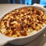A close-up of a hot, bubbly bacon popper dip baked to golden perfection in a white oval casserole dish.