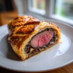 A cross-section slice of beef wellington recipe showing medium-rare beef tenderloin wrapped in golden puff pastry and duxelles.