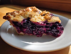 A close-up of a slice of homemade blueberry pie recipe showing juicy, dark purple filling and a golden, flaky crust.