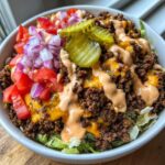 A close-up of a cheeseburger bowl featuring seasoned ground beef, melted cheese, special sauce, pickles, tomatoes, and onions over lettuce.