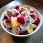 Close-up of a white bowl filled with cheesecake fruit salad featuring strawberries, blueberries, pineapple, and grapes mixed in a creamy topping.