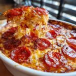 A spoonful of hot, cheesy pepperoni pizza dip is lifted, showing long, stretchy cheese pulls.