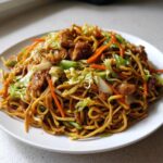 A close-up of a serving of homemade chicken chow mein recipe tossed with noodles, cabbage, and carrots on a white plate.
