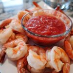 Close-up of cooked shrimp arranged around a small glass bowl of red cocktail sauce for shrimp cocktail.