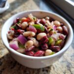A close-up of a vibrant, mixed bean salad featuring white beans, kidney beans, red onion, and green pepper in a white bowl.