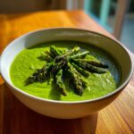 A vibrant green bowl of creamy asparagus soup garnished with roasted asparagus spears.