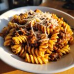 Close-up of a white bowl filled with creamy beef pasta (rotini shape) topped generously with shredded Parmesan cheese.