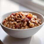 A white bowl filled with creamy black eyed peas topped with crispy bacon pieces.