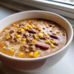 A close-up shot of a hearty bowl of cowboy soup featuring ground beef, bright yellow corn, and dark red kidney beans in a creamy broth.