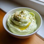 A close-up of a light green, whipped olive dip swirled in a white bowl, drizzled with olive oil and sprinkled with spices.