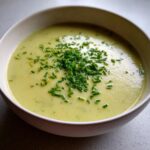 A close-up of creamy, pale green leek soup garnished generously with bright green chopped chives in a white bowl.