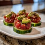 Four servings of tuna tartare stacked on thick cucumber slices, topped with avocado and sesame seeds.