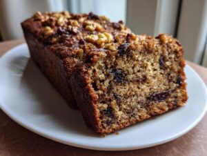 A thick slice of moist date nut bread topped with walnuts, resting next to the loaf on a white plate.