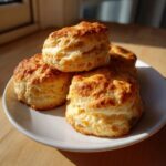 Four golden brown, flaky homemade scones stacked on a white plate, highlighted by sunlight.