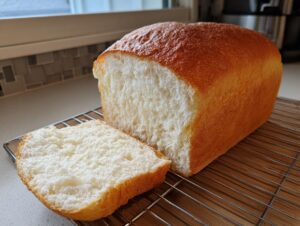 A freshly baked loaf of Japanese milk bread, golden brown on the crust, with one fluffy slice resting beside it on a cooling rack.