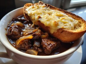 Close-up of a bowl of rich french onion beef stew topped with a thick slice of bread covered in melted, browned cheese.